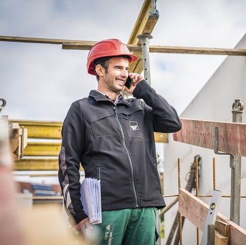 ein Mann traegt weisenburger Arbeitskleidung und einen roten Bauhelm, er steht laechelnd auf einer Baustelle und telefoniert