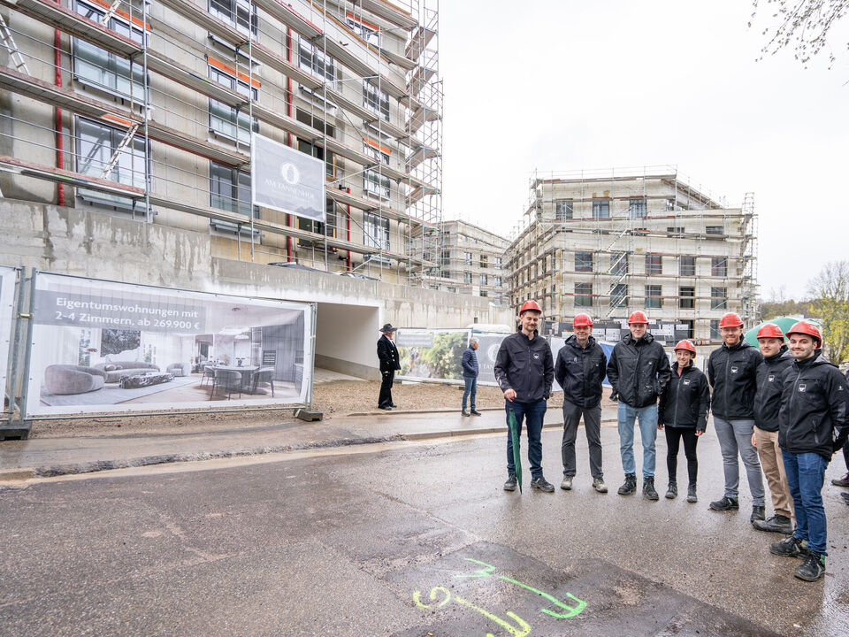 Gruppe von Menschen mit rotem Bauhelm stehen vor einer Baustelle 