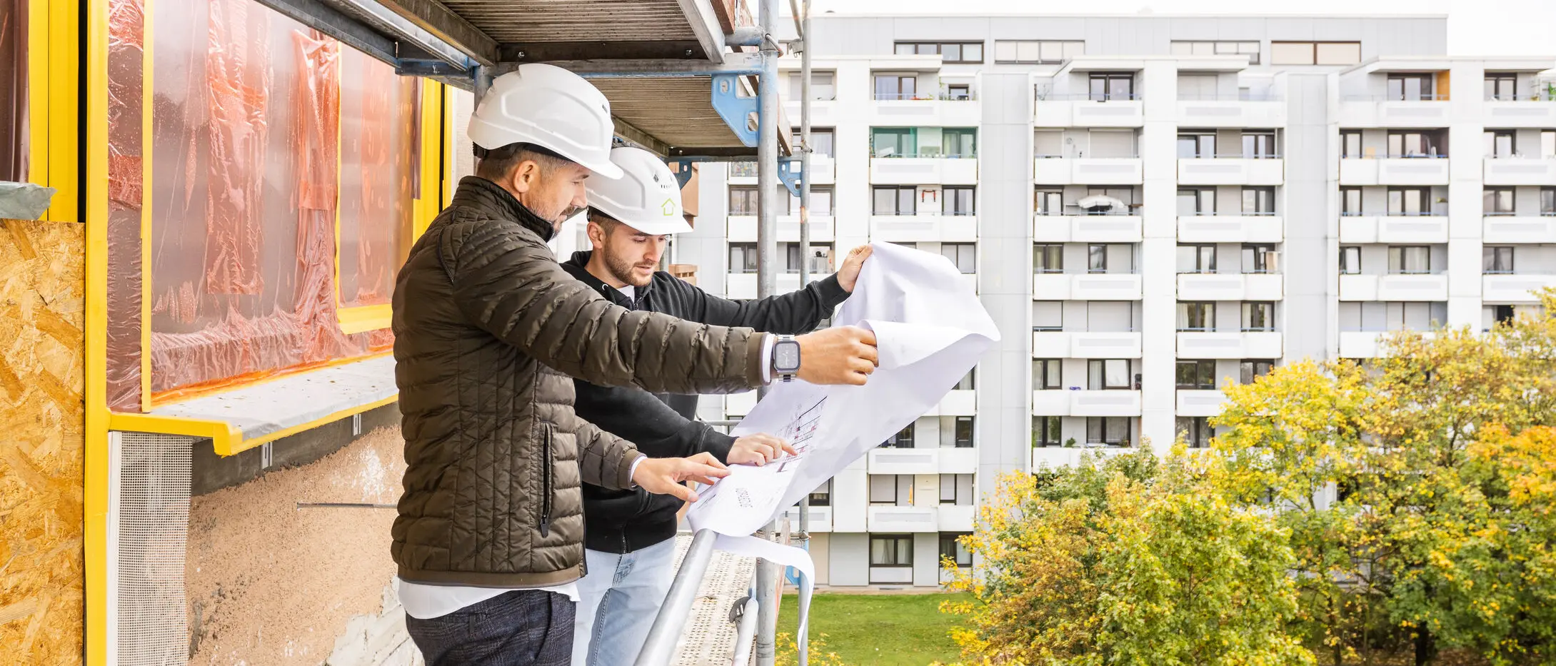 sanierungsprofi team at refurbishment of apartment buildings Two men wearing construction helmets stand on scaffolding and look at a 2D construction plan
