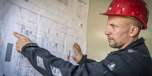 Mann mit Bauhelm schaut sich ein 2D Architekturplan an und zeigt auf diesen 