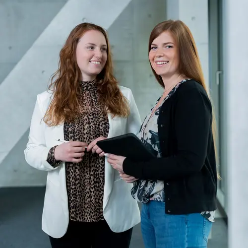 Der Bereich Projektentwicklung bei weisenburger Eine junge Frau hält einen Stift in der Hand und lächelt eine junge Frau an die ein Tablet in der Hand hat
