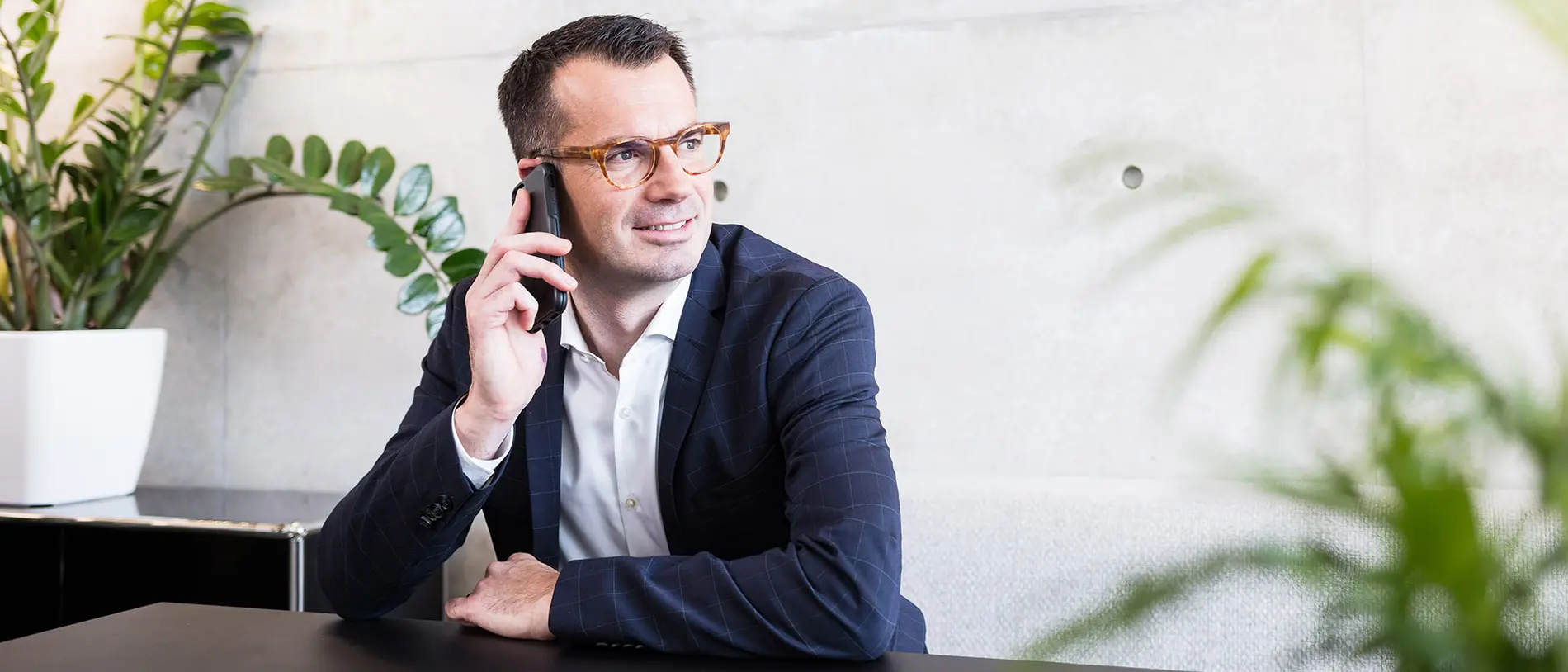 Kontaktbanner A man with glasses is sitting at a table in front of a houseplant and is talking on the mobile phone