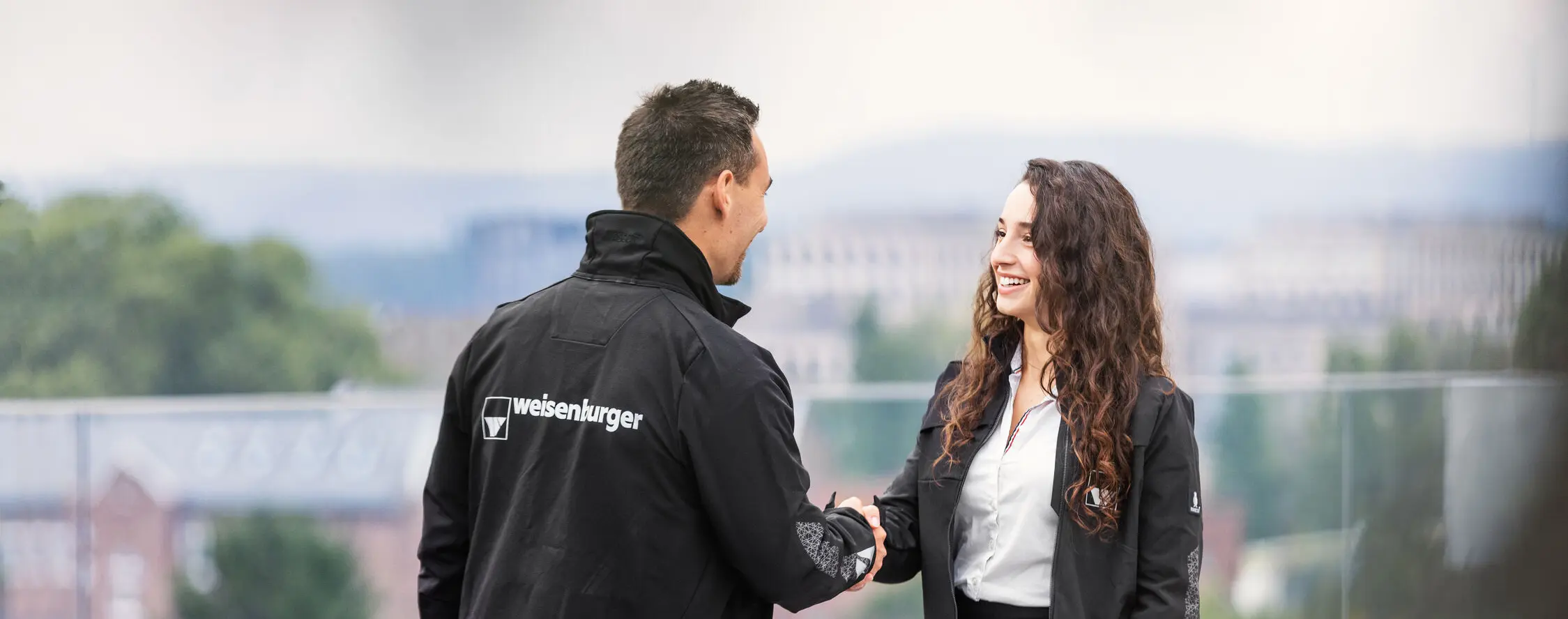 weisenburger-Mitarbeiter mit Handschlag Ein Mann und eine Frau in weisenburger Jacken schütteln sich gegenseitig die Hände auf der weisenburger Dachterrasse