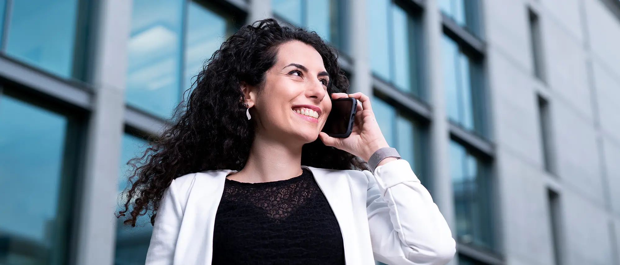 Kontakt Banner Eine weisenburger-Mitarbeiterin mit schwarzen Locken telefoniert laechelnd vor einem modernen Gebaeude