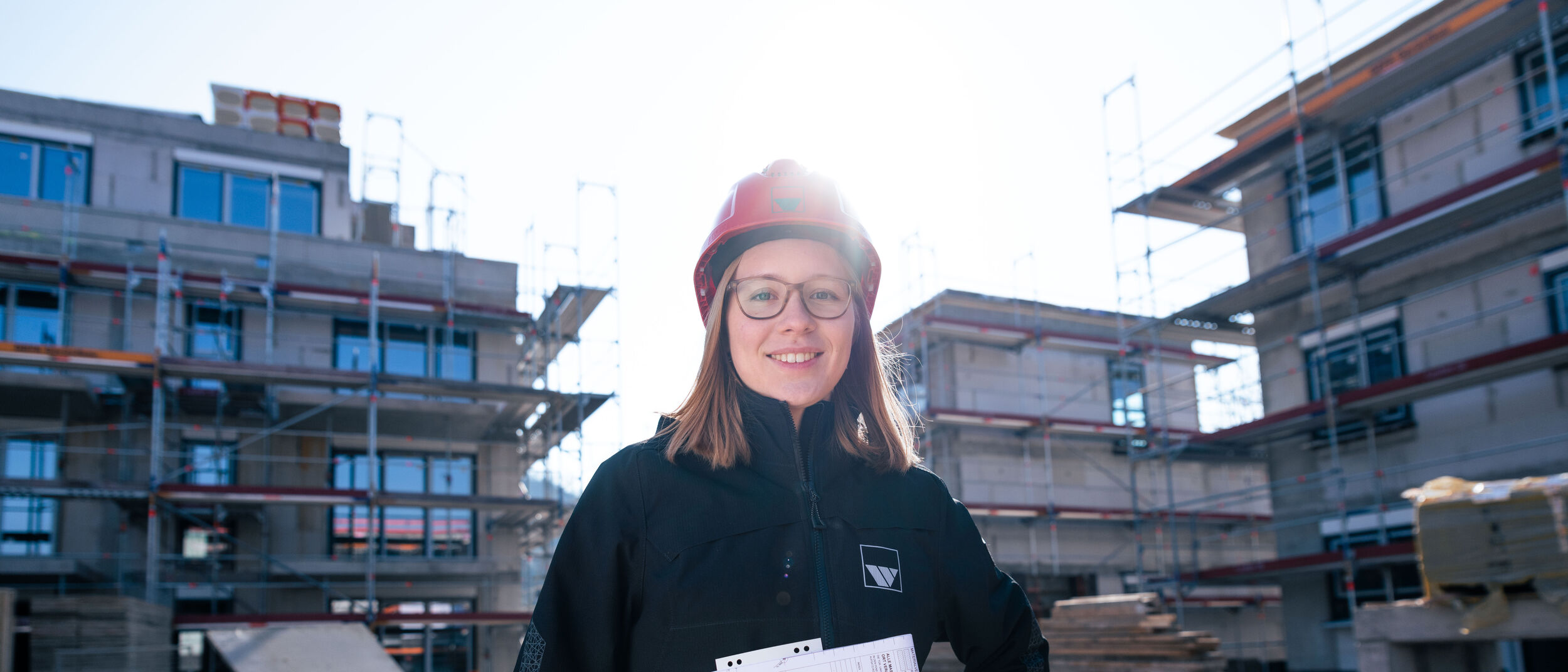 eine junge Frau mit rotem Bauhelm und einer weisenburger Jacke steht auf einer Baustelle und laechelt 