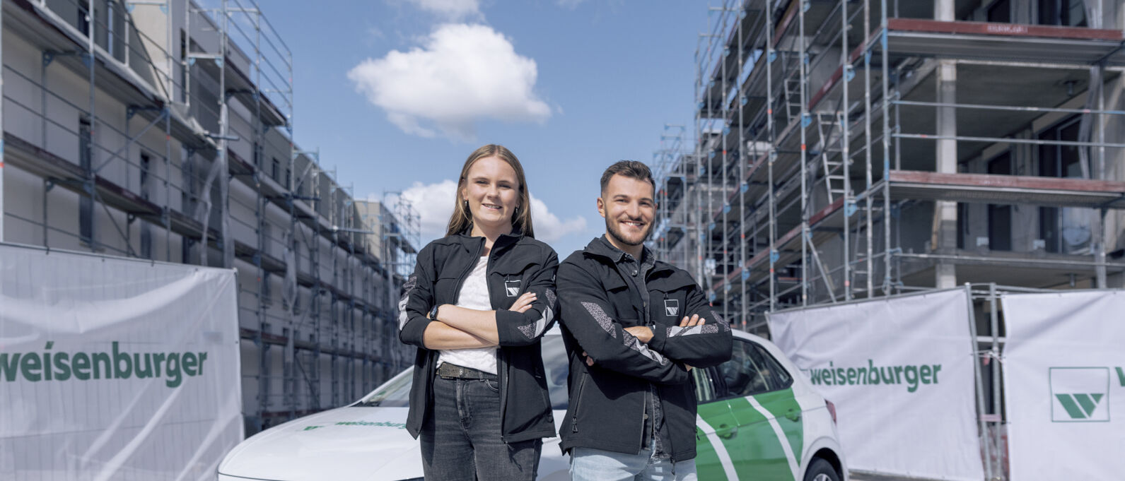 eine junge Frau und ein junger Mann verschraenken beide die Arme und laecheln, sie stehen vor einem weisenburger Auto auf einen Baustelle  