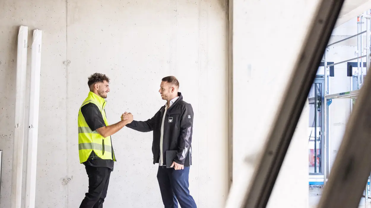 Ein Berufseinsteiger und der Ausbilder sind auf einer weisenburger Baustelle Zwei Männer mit weisenburger Jacken begrüßen sich gegenseitig mit einem Handschlag