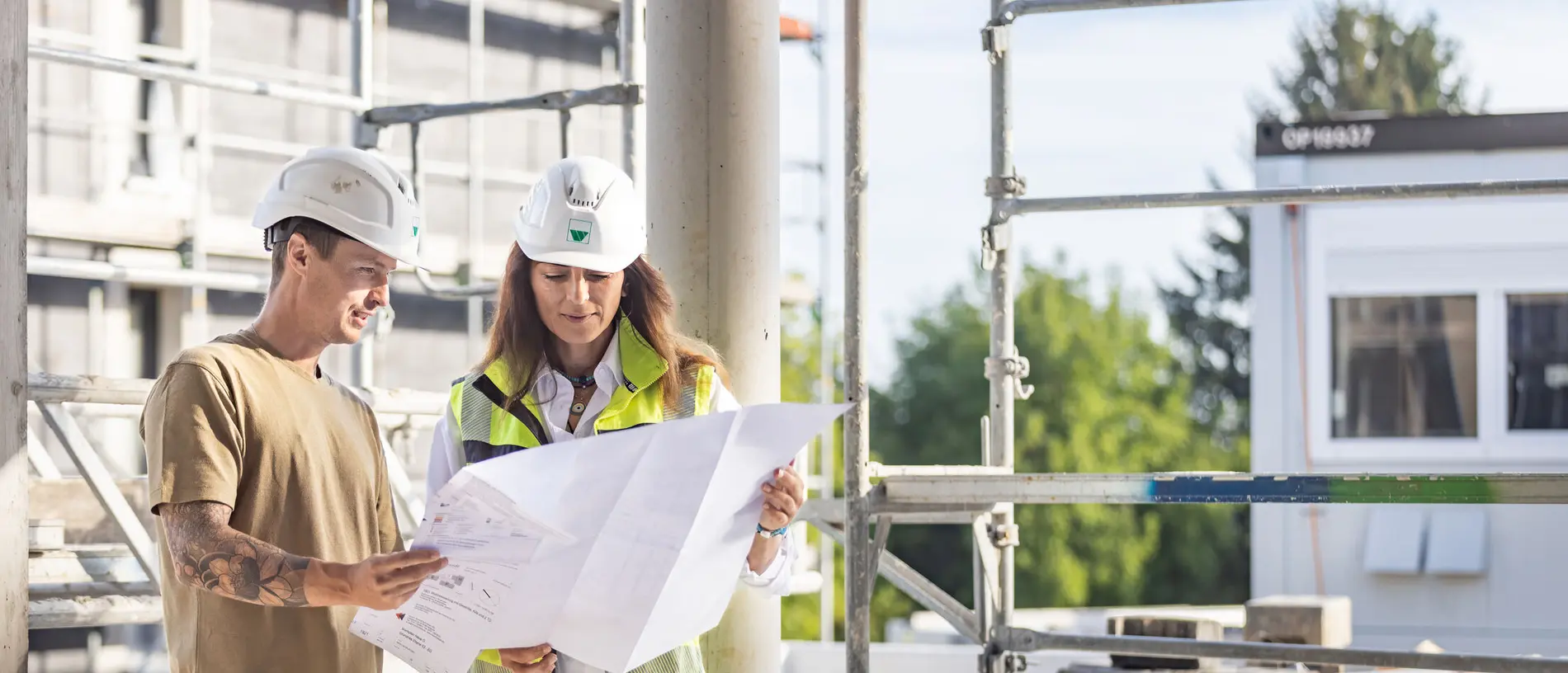 weisenburger-Mitarbeiter bei der Bauplanung auf der Baustelle ein Mann und eine Frau tragen beide einen Bauhelm und schauen einen Bauplan an, hinter ihnen ist ein Gerüst aufgestellt
