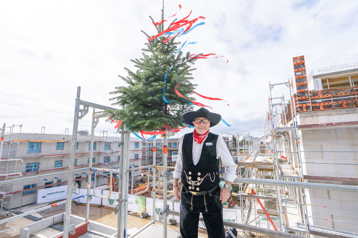 Ein Mann in traditioneller Handwerkskleidung steht auf einem Baugerüst vor einem Richtbaum