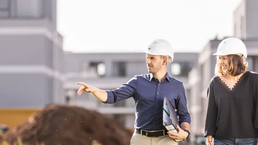 weisenburger-Bauleitung bei der Baustellenbesichtigung Ein Mann und eine Frau mit Bauhelmen laufen lächelnd über eine Baustelle, der Mann hält in seiner Hand ein Ordner