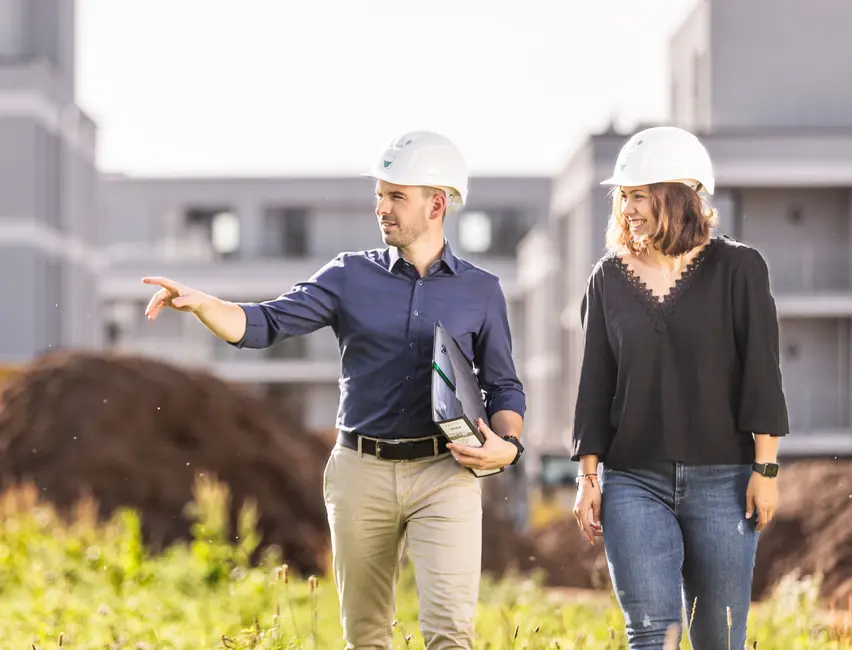 weisenburger-Bauleitung bei der Baustellenbesichtigung Ein Mann und eine Frau mit Bauhelmen laufen lächelnd über eine Baustelle, der Mann hält in seiner Hand ein Ordner