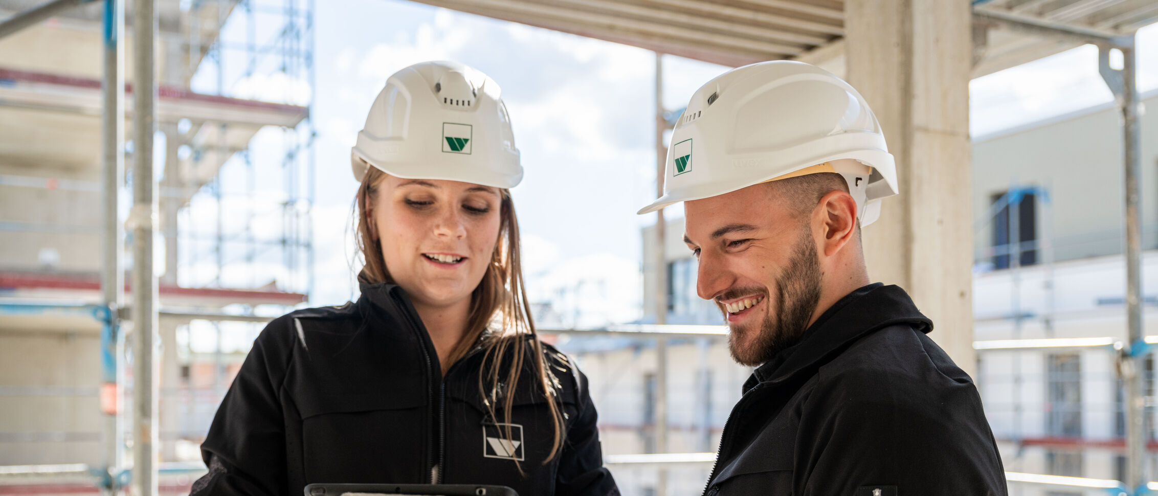 eine junge Frau und ein junger Mann, mit weißen Bauhelmen, stehen auf einer Baustelle und schauen gemeinsam auf ein Tablet 