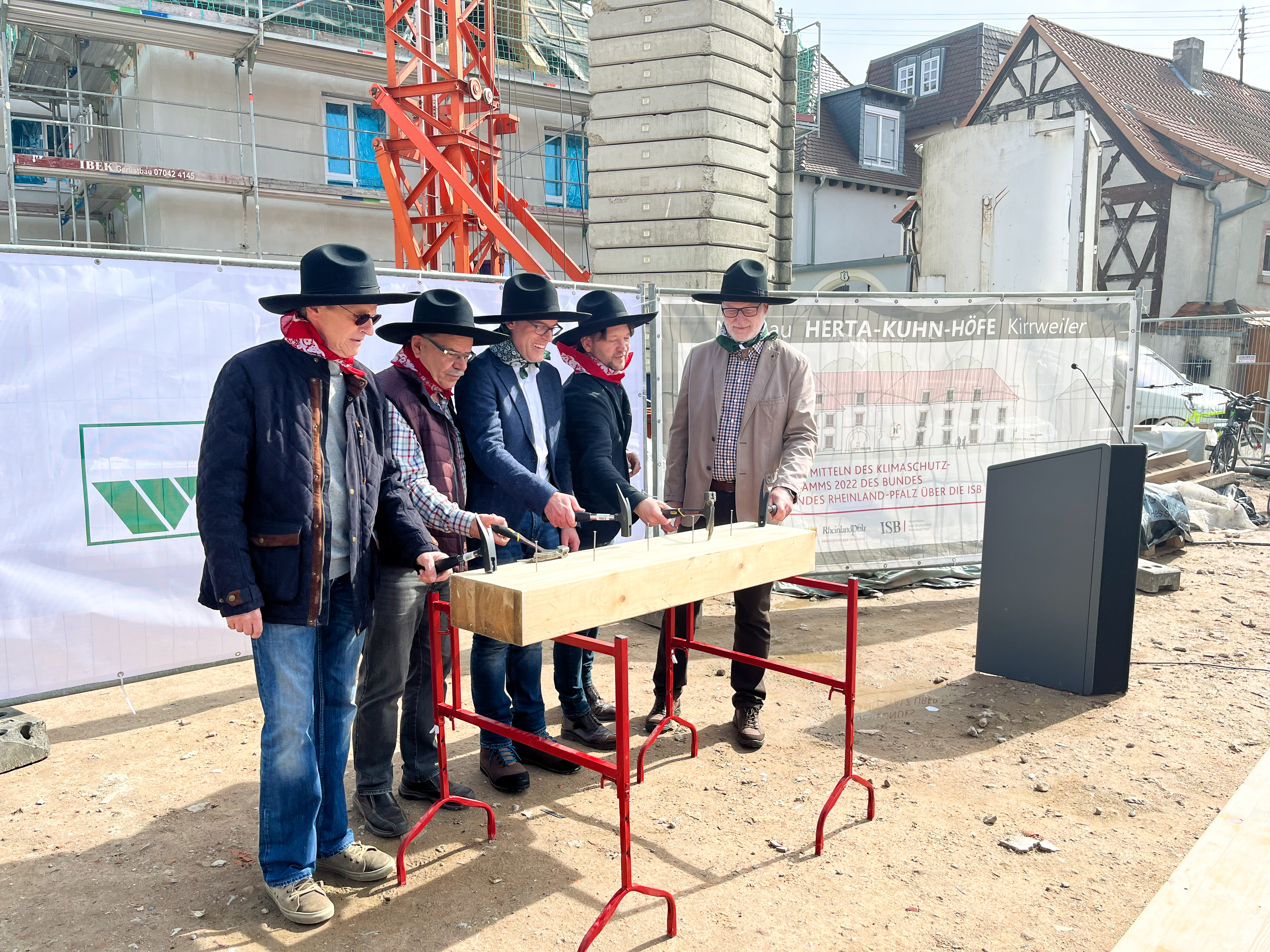 Fünf Männer mit schwarzem Hut stehen auf einer Baustelle und schlagen jeweils einen Nagel in einen Holzblock