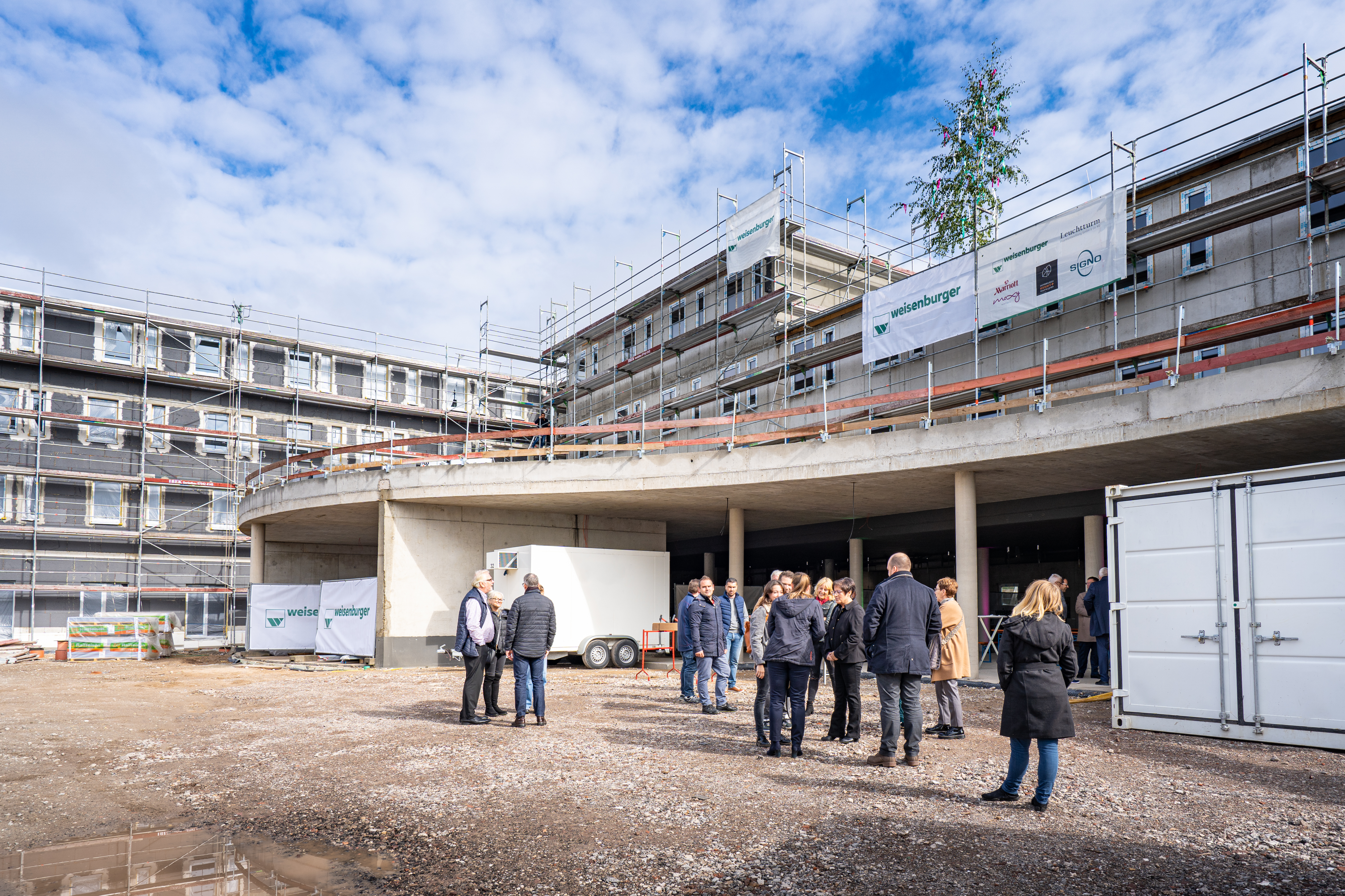 Eine Baustelle mit einem halbfertigen Gebäude und einer Gruppe von Menschen davor.