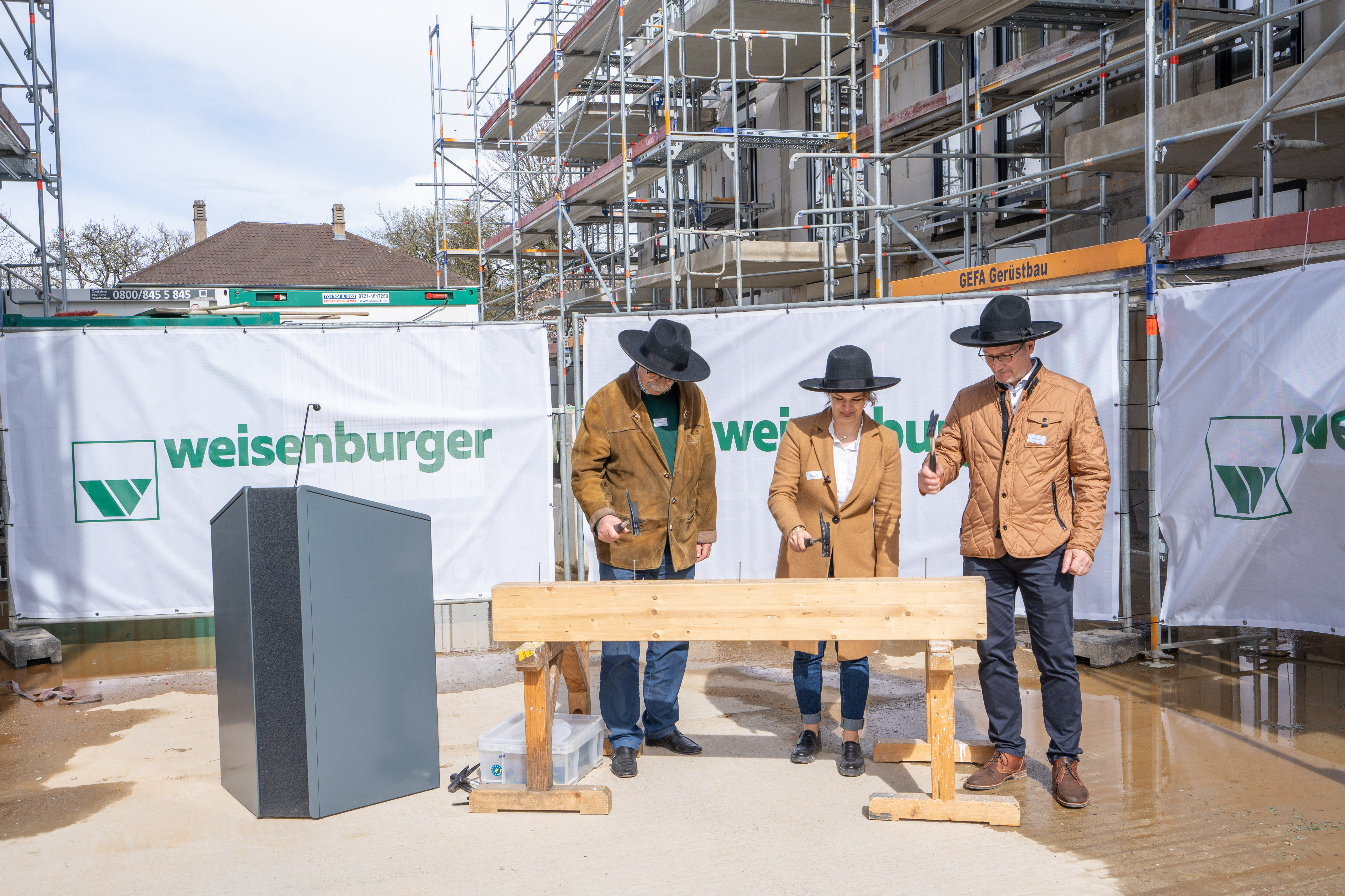 Drei Personen in Handwerkskleidung stehen auf einer Baustelle vor einem Holzbalken mit "weisenburger"-Banner im Hintergrund