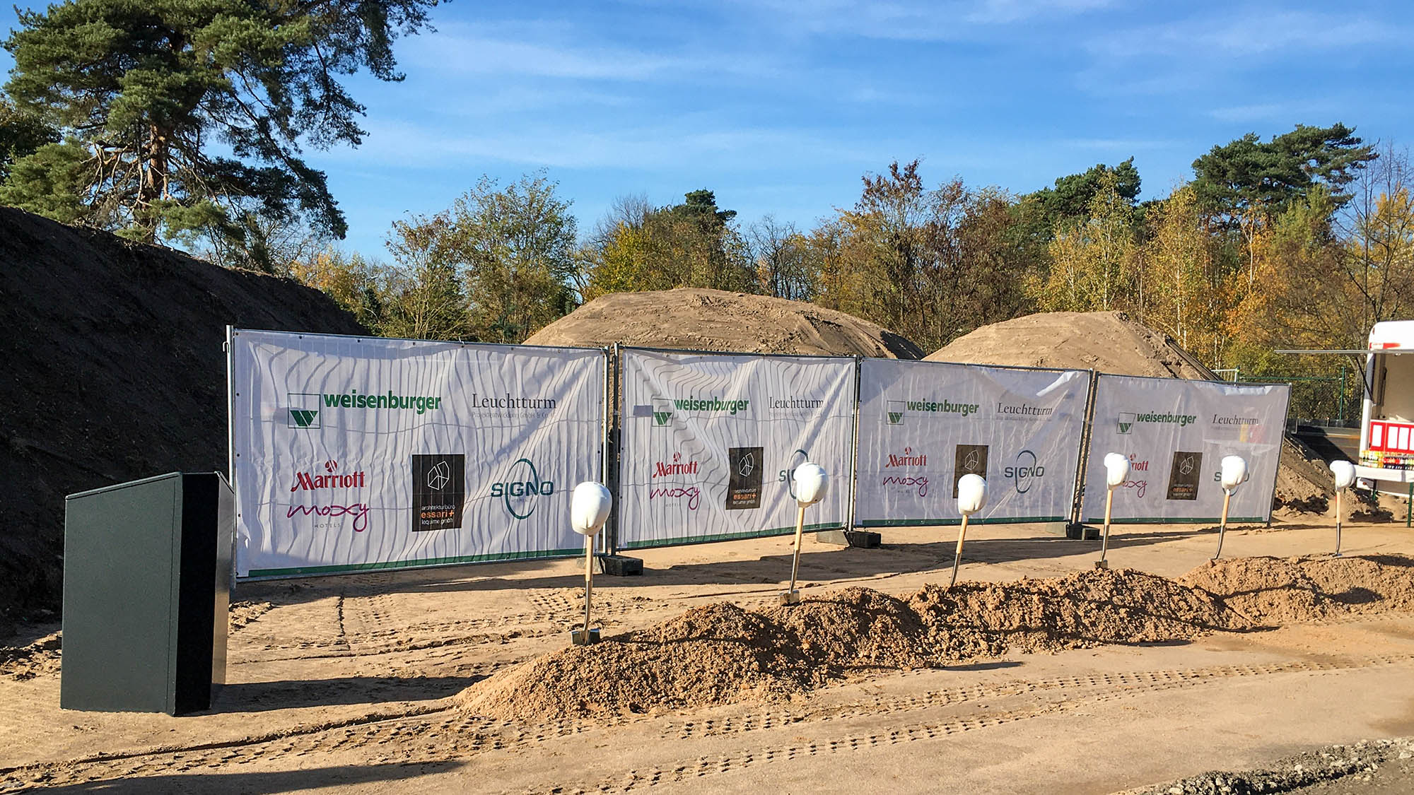  Groundbreaking: Several spades are stuck in an elongated pile of earth, behind them tarpaulin with the logos of the companies involved