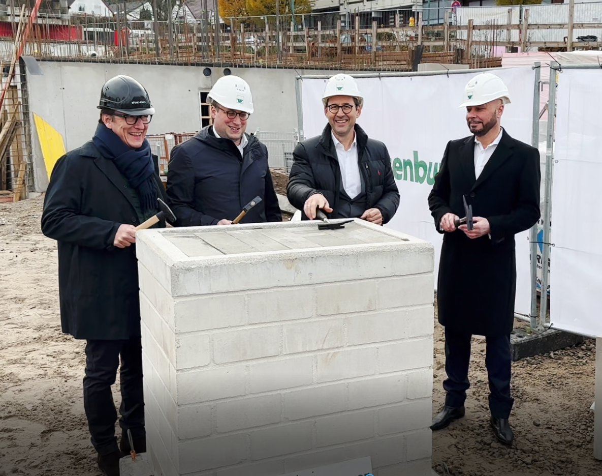 Vier Männer mit Bauhelmen stehen auf einer Baustelle und halten Hammer in der Hand, während sie an einer Grundsteinlegung teilnehmen